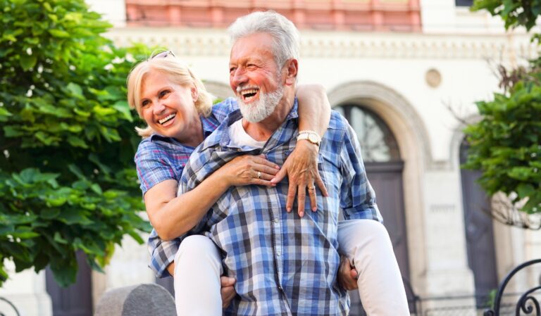 Older couple enjoying a playful moment outdoors; woman sits on the man's shoulders, both laughing.