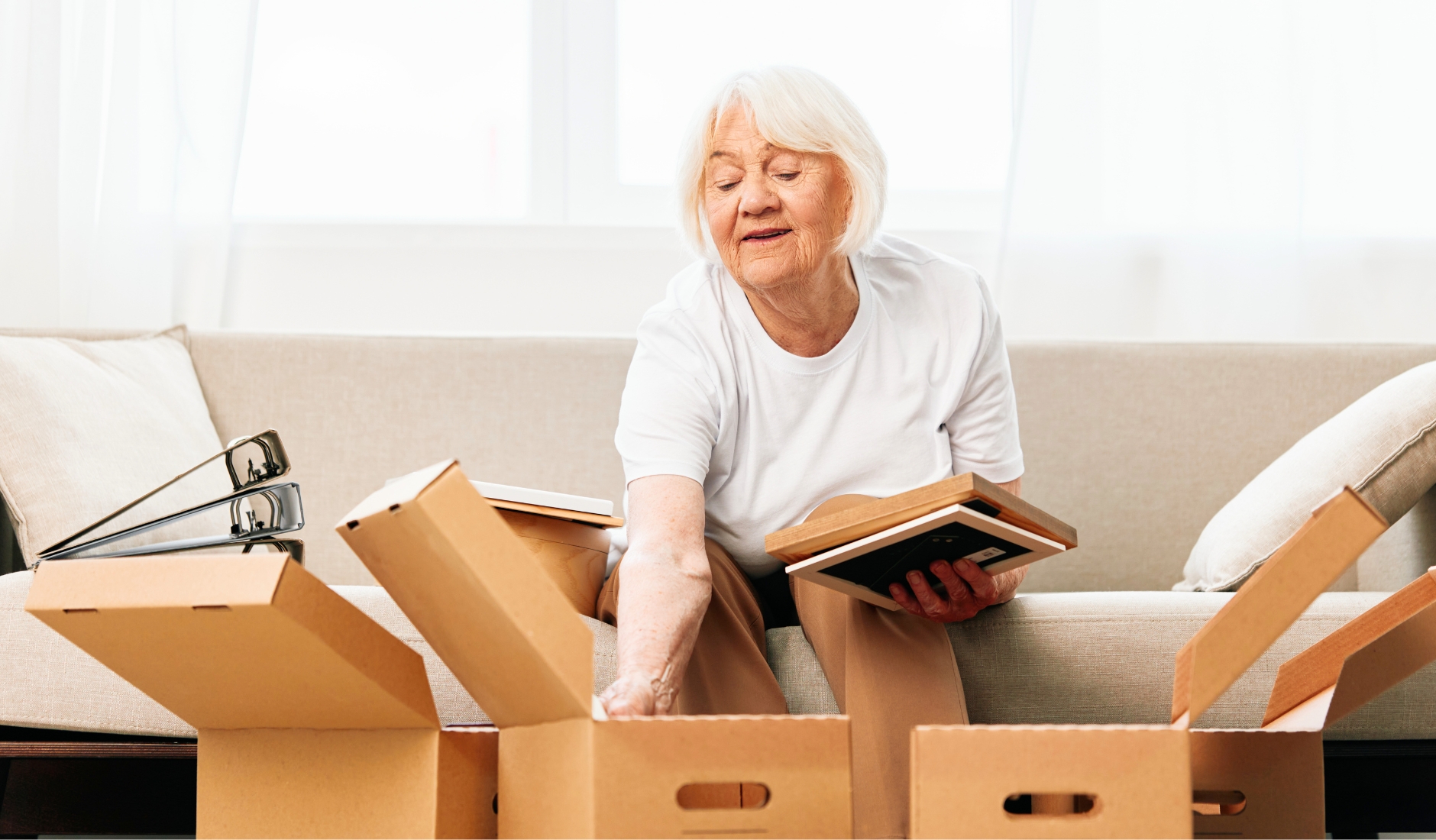 Older woman sits on a beige sofa, unpacking moving boxes and handling a photo album or book near to her.